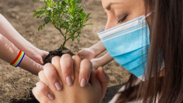 A person wearing a blue surgical mask is seen in profile, with their eyes closed in contemplation. In the foreground, several hands of diverse individuals are shown working together to plant a small green plant in dark soil. There is a hint of a rainbow bracelet on one of the hands, symbolizing unity and diversity.