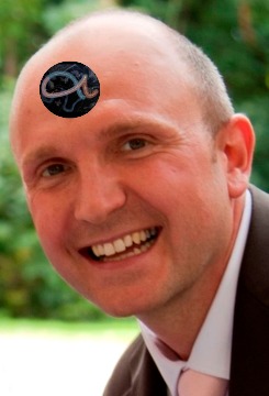 A cheerful bald man in a suit, smiling broadly. He has a symbolic depiction of Pisces from the Mazzaroth overlaid on his forehead.