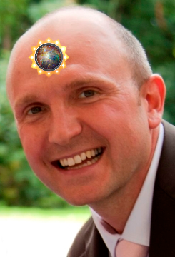Ein kahlköpfiger Mann mit einer hellen, strahlenden Himmelsgrafik, die einen Teil des Mazzaroth darstellt, mittig auf seiner Stirn, lächelt breit und trägt einen dunklen Anzug und eine violette Krawatte.