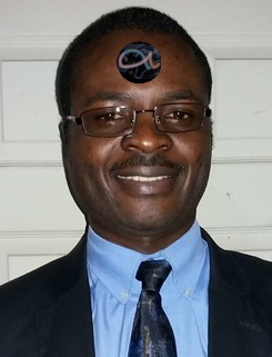 A smiling man wearing glasses and dressed in a blue shirt with a dark tie, with a symbol reminiscent of a Mazzaroth constellation on his forehead.