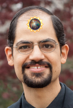 A smiling man with glasses has a symbolic circular emblem depicting a cosmic element positioned on his forehead, all set against a blurred natural background.