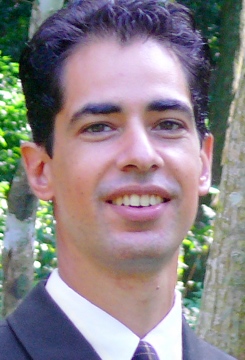 A portrait of a smiling man in a dark suit and white shirt, with neatly styled black hair, standing outdoors with green foliage in the background.