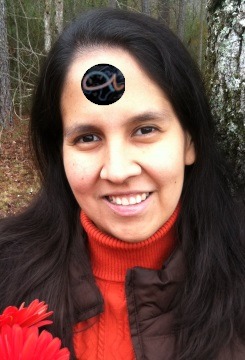 A smiling woman with long dark hair, wearing an orange turtleneck and a brown jacket, holding red flowers. She has a symbol related to the Mazzaroth placed on her forehead.