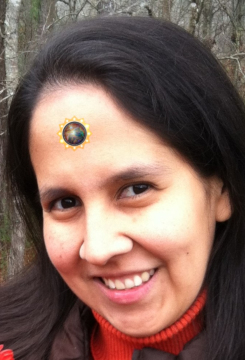Una mujer de cabello largo y oscuro, con una sonrisa radiante y un suéter rojo de cuello alto, posa al aire libre. Lleva un emblema circular con un sol brillante en la frente.