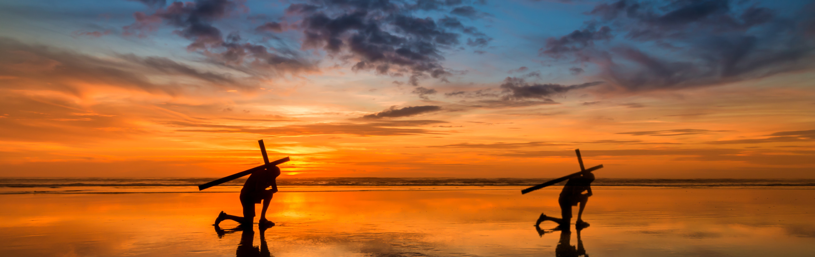 Silhouetted figures carrying surfboards walk along a beach at sunset, with a vivid display of orange and blue hues reflected on the wet sand, evoking the vast canvas of the heavens at the close of day.