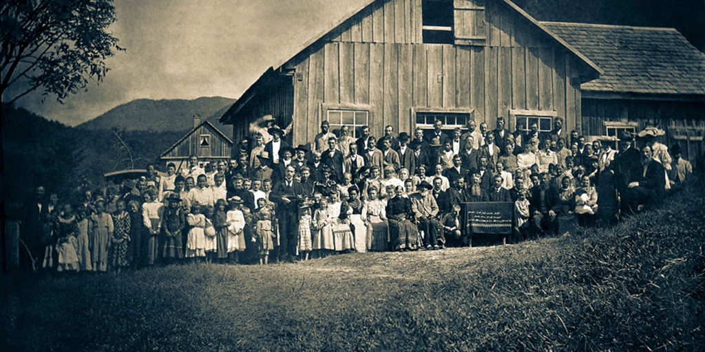 A historical photograph showing a large group of people gathered in front of a rustic wooden building, surrounded by lush countryside. The scene captures a mix of adults and children, some seated on benches, others standing, with a backdrop of rolling hills and a clear sky.