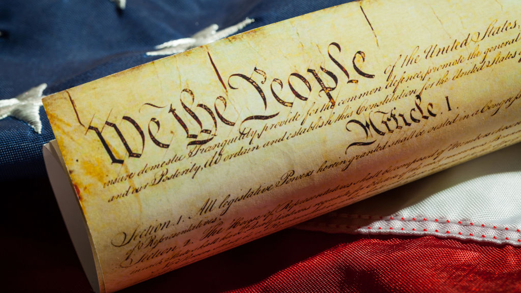 Close-up of an aged parchment bearing the ornate, handwritten script "We the People" reflecting its historical significance, nestled against a background comprising part of the American flag, displaying stripes of red and white.