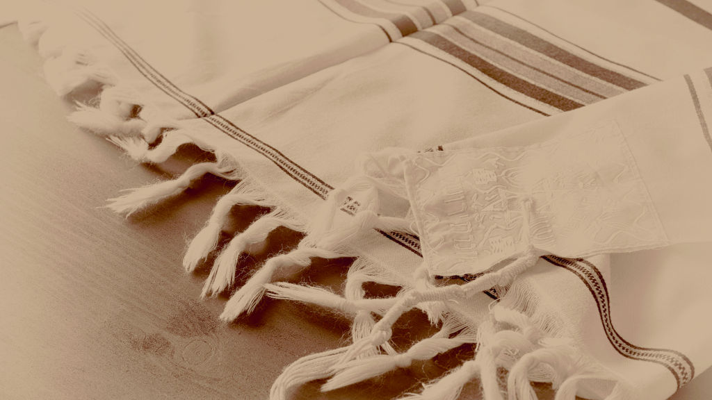Sepia-toned image of a Jewish prayer shawl, known as a Tallit, with intricately embroidered symbols and fringed edges laid on a flat surface.