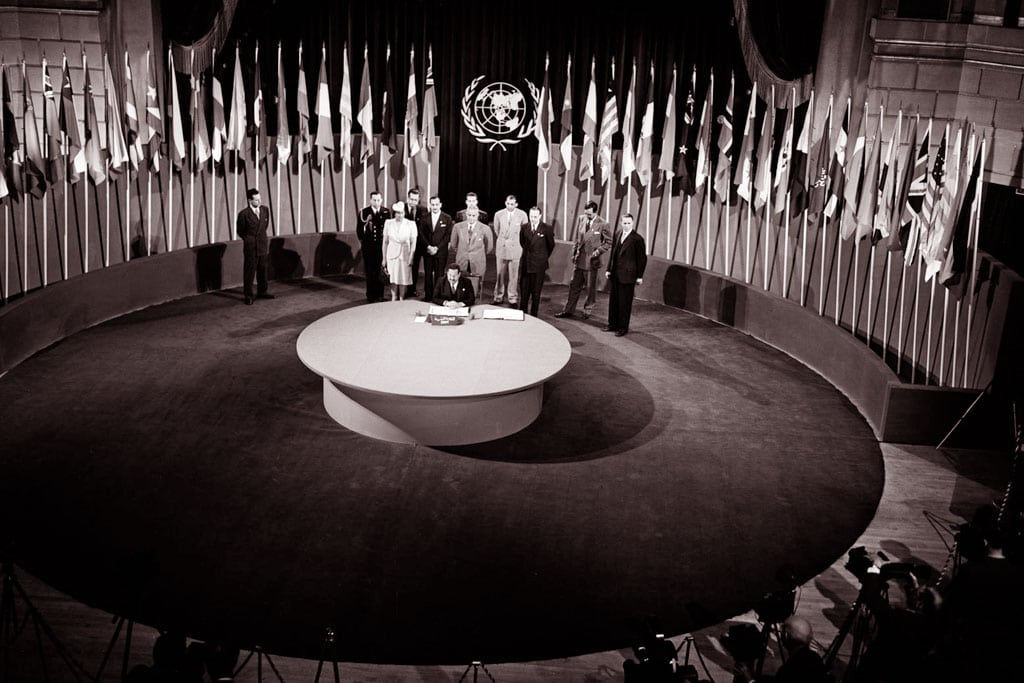Black and white photograph of a historic international gathering inside a large hall with numerous flags representing various countries. A group of dignitaries is standing on a circular platform beneath a large emblem of the United Nations.