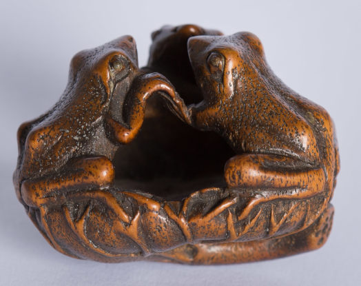 A wooden carving depicting two frogs sitting opposite each other, their mouths open as if in conversation, set against a neutral background. The sculpture exudes a rustic charm, highlighted by its detailed textures and the evident craftsmanship.