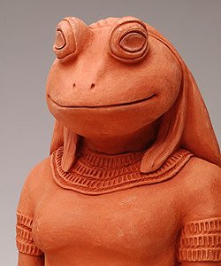 A terracotta sculpture of a whimsical frog standing upright, depicted with a human-like posture and wearing a detailed textured sweater. The sculpture features large, circular eyes and a subtle, content smile.