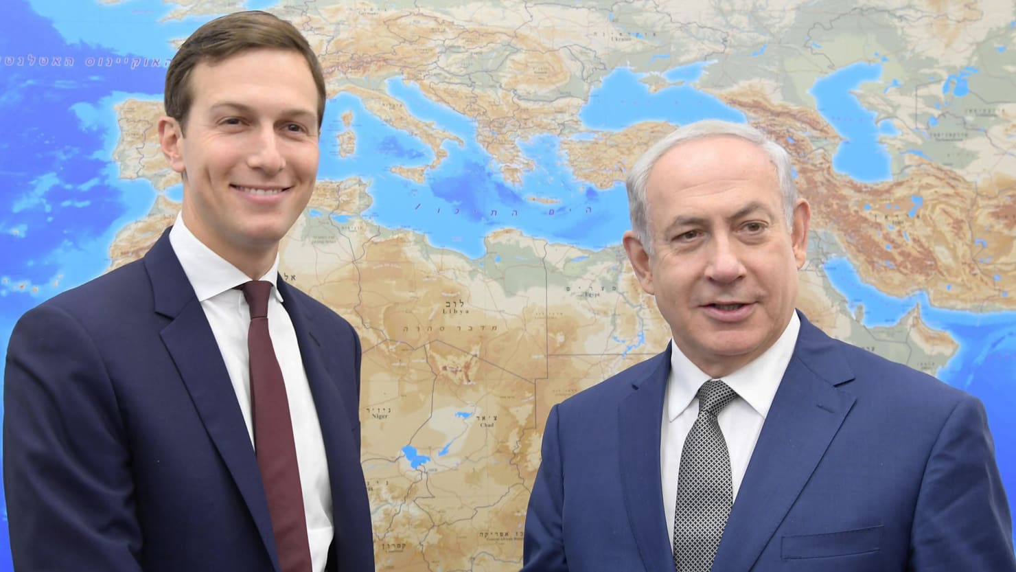 Two men in business suits, smiling, stand in front of a large wall map displaying various regions, primarily highlighting the Mediterranean area. Their poised appearance and the displayed map suggest a context of geographical or political discourse.