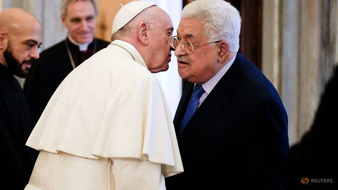 Pope Francis and President Mahmoud Abbas share a close greeting, their foreheads nearly touching, in a warmly lit hall with other dignitaries in the background.