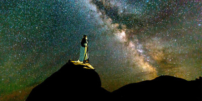 A person stands on a mountain top at night, gazing at a vast star-filled sky including the Milky Way's dense star clusters and dust lanes.