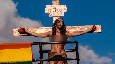 A person is portrayed in a tableau resembling a crucifixion, with arms extended and attached to a wooden cross bearing the inscription "Homophobia LGBT." They appear bloodstained and wear a crown of thorns, standing above a rainbow flag under a partly cloudy sky.