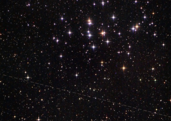 A vast night sky filled with numerous twinkling stars varying in color and intensity. A distinct trail of a satellite or a piece of space debris creates a linear streak across the star-filled backdrop.