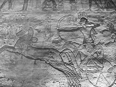 Ancient bas-relief carving depicting a human figure with a drawn bow, riding a chariot pulled by galloping horses. The art also features symbolic diagrams, including circles and patterns potentially representing celestial bodies and their orbits.
