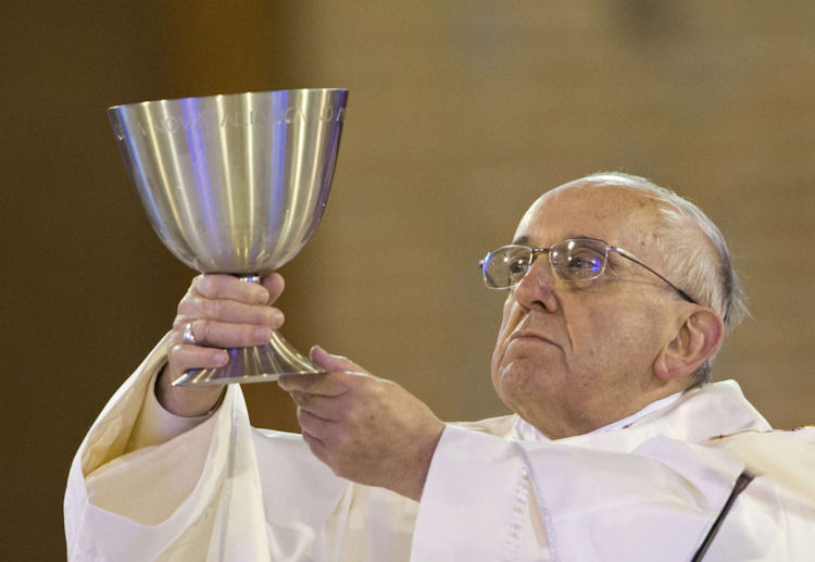 An elderly man in white religious vestments raises a large silver chalice with both hands during a ceremony. His expression is contemplative and focused.