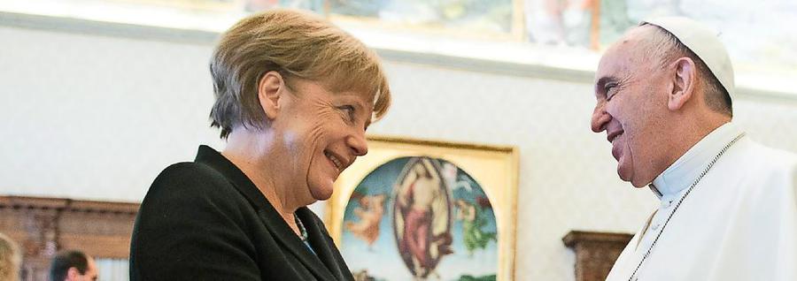 A woman and a man, both smiling, engaged in a conversation. The woman, dressed in a dark suit, is on the left while the man, wearing religious attire, stands on the right. In the background, a painting depicting a biblical scene is visible.