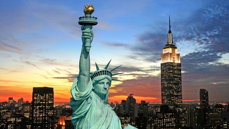 An image showing the Statue of Liberty on the left with its torch held high against a vibrant twilight sky, and the Empire State Building on the right, illuminated at its spire. The backdrop features a sunset over a cityscape with skyscrapers silhouetted against the fading light.