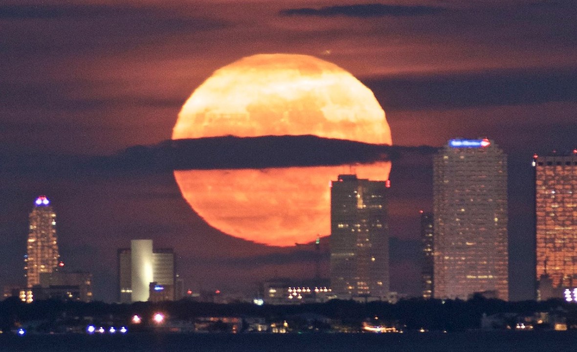 A large, radiant full moon, glowing orange, rises behind the silhouette of a city skyline under a dusky sky. A cloud bisects the moon creating a dramatic visual contrast.