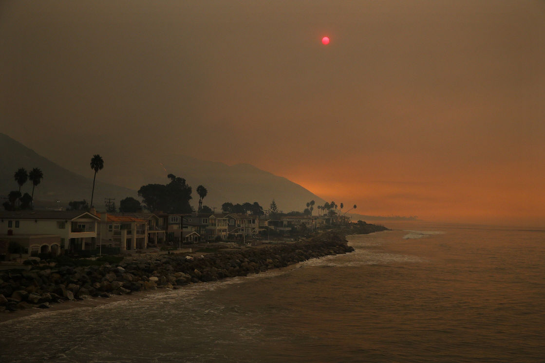 A coastal scene under a smoky, orange sky with a visibly dimmed red sun, casting a surreal glow over a line of houses and palm trees along a rocky shoreline. Mountains faintly visible in the background contribute to the ethereal atmosphere.