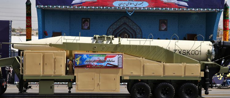 A military parade featuring a large ballistic missile mounted on a multi-axle launch vehicle. The vehicle is parked in front of a platform with traditional architectural designs and religious banners.