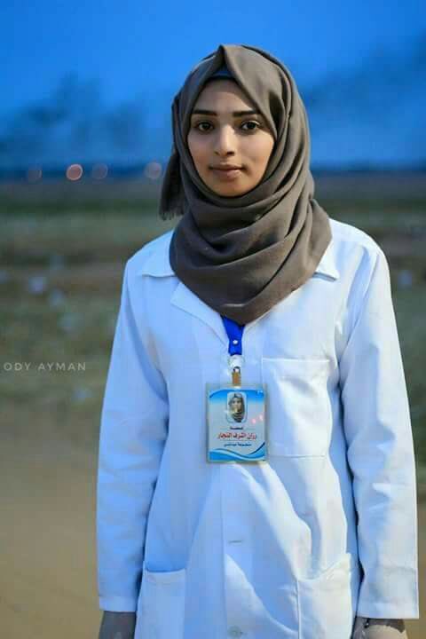 A young woman with a confident expression stands outdoors during twilight, wearing a white lab coat and a brown hijab. She has an ID badge clipped to her coat displaying her name and photo. The backdrop features a blurred early evening sky and hints of the first visible stars.