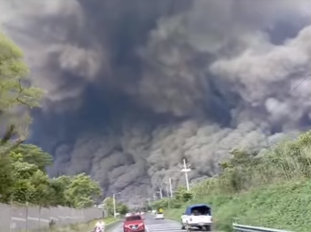 A dramatic scene as vehicles on a highway flee from a massive, rapidly advancing pyroclastic flow from a volcanic eruption, with a towering, dense ash cloud engulfing the sky above.