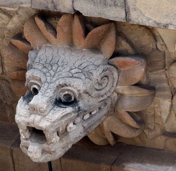 Stone sculpture of a mythical creature's head mounted on a wall, featuring large, bulging eyes, curled horns, and a row of pointed teeth. The creature is surrounded by stylized stone carvings resembling celestial rays.