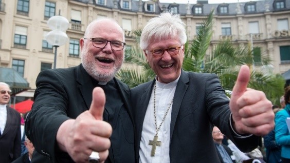 Two joyful clergy members, one wearing glasses and a black suit, and the other wearing a white cross necklace and a black suit, standing outdoors with their thumbs up, smiling broadly in a crowded public square.