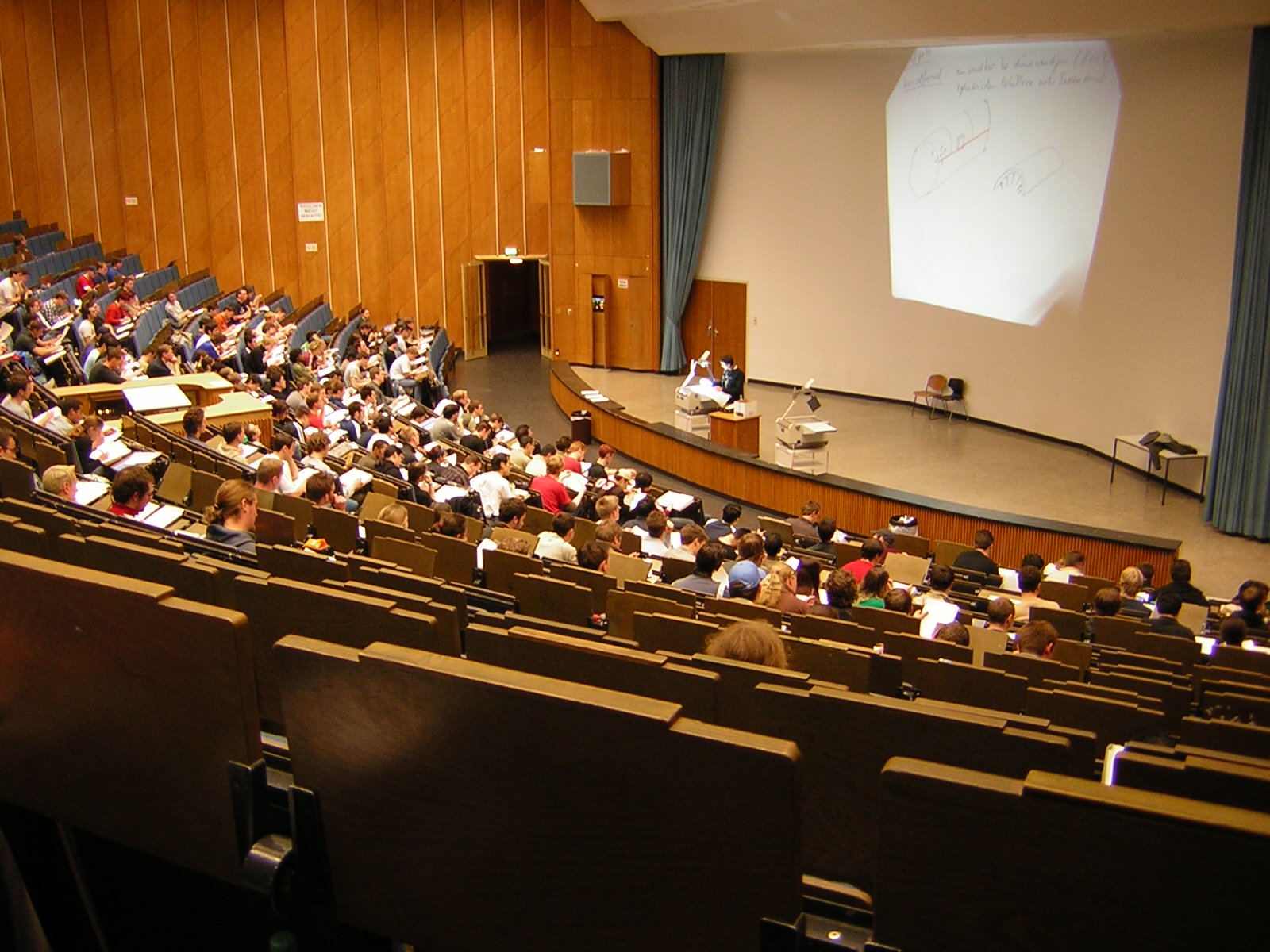 A large university lecture hall filled with attentive students focusing on a presentation. The lecturer at the front uses a projection screen displaying diagrams related to celestial bodies and their dynamics, avoiding references to the Mazzaroth.