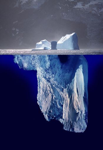 An iceberg is shown with a majority of its mass submerged underwater, demonstrating its vast size beneath the sea surface compared to the smaller visible portion above. Background gradient ranges from near white to deep blue, contrasting the iceberg with the surrounding waters.