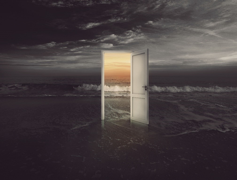 An open door stands isolated on a sandy beach during twilight, with waves gently crashing near its threshold. Through the door frame, a radiant sunrise is visible, casting a warm glow.
