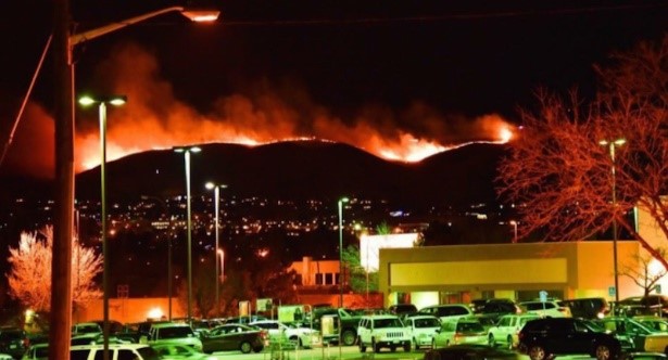 Nighttime view of a city landscape with a parking lot full of cars in the foreground. In the background, large wildfires light up the hills, casting a dramatic orange glow in the sky, contrasted by the urban lights below.