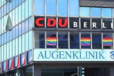 Modern building facade featuring a sign that reads "CDU BERLIN AUGENKLINIK" in block letters, with three flags designed with a spectrum of colors flying beneath the sign.