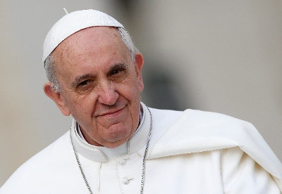 Portrait of an elderly man wearing a white skullcap and religious attire, smiling gently against a blurred background.
