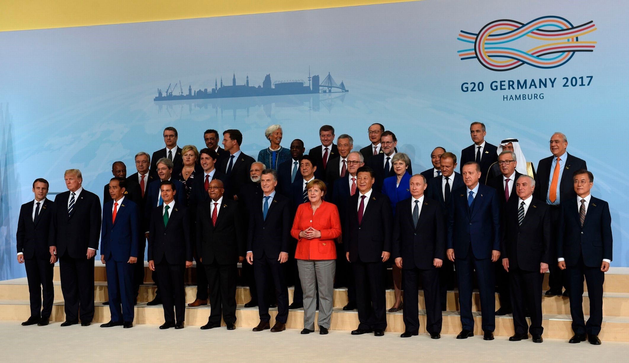 Group photo of global leaders at the G20 Germany 2017 summit in Hamburg. Leaders from various nations are standing together, posed in three staggered rows on a stage, with a background depicting a stylized cityscape and a large logo reading "G20 Germany 2017 Hamburg."