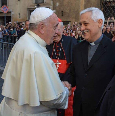 Pope Francis smiling and shaking hands with a priest in a casual outdoor setting, accompanied by a cardinal in the background.