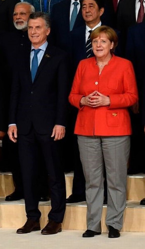 A formal group photo featuring two prominent figures in the foreground. On the left, a man in a dark suit and blue tie stands with a pleasant expression. Next to him, a woman in a red jacket and grey trousers smiles gently, her hands clasped in front. Several other formally dressed individuals are partially visible in the background.