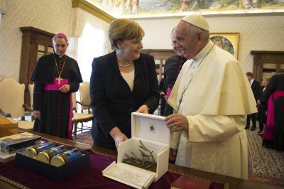 Chancellor Angela Merkel and Pope Francis engage in a warm conversation while exchanging gifts in an ornately decorated room. Merkel is presenting a framed piece to the Pope, who is dressed in traditional papal attire.