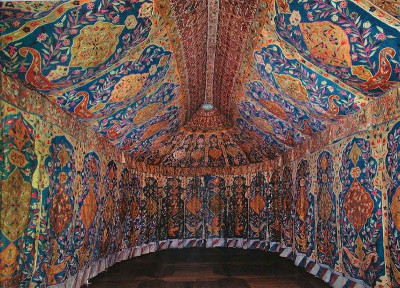 Interior view of a vibrantly decorated dome, featuring elaborate mosaic depicting various celestial bodies and human figures, possibly portraying the Mazzaroth set against a night sky. The artwork includes intricate patterns and bright, earthy colors, enveloping the ceiling and arches of the room.