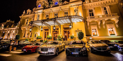Nighttime view of a grand illuminated building with classical architecture, featuring ornate facades and balconies, with a line of luxury vehicles parked in front.