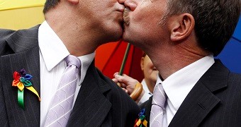 Two men in formal attire kissing, each wearing a rainbow ribbon on their lapels, with a multicolored background.