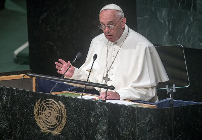 A religious leader in white attire is speaking at a podium with the emblem of the United Nations in the background, gesturing with his right hand.