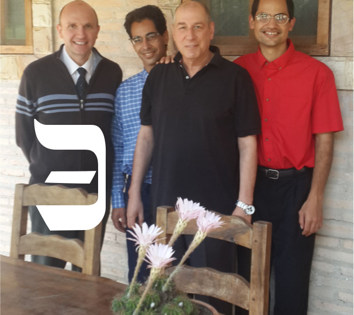 Four men smiling at the camera, standing behind a wooden table with a centerpiece of pink flowers. The first man is in a dark blue sweater with horizontal white stripes, the second in a blue shirt, the third in a black shirt, and the fourth in a bright red shirt.