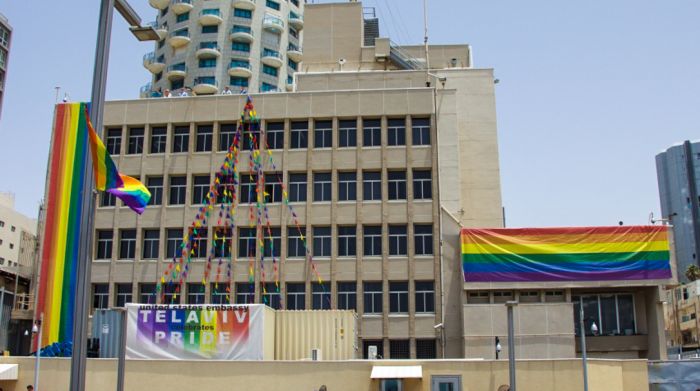 Photo of a cityscape featuring a modern building adorned with multiple rainbow flags and a decorative structure resembling an abstract pyramid composed of colorful triangular flags. A banner reads "Tel Aviv Celebrates Pride".