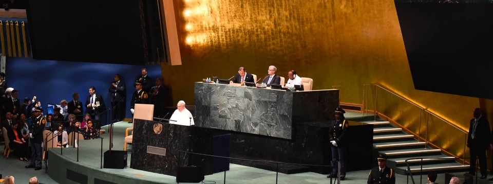 Pope Francis addressing delegates at a United Nations assembly, standing at a podium in a large hall with an audience and dignitaries seated and standing around, beneath a large gold backdrop.