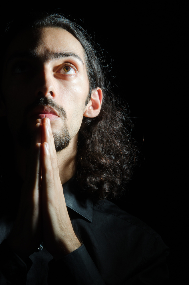 A man with long, curly dark hair and a beard is depicted in a contemplative pose, his fingers pressed together in prayer against his chin. He looks upwards with a focused gaze, illuminated by a soft, directional light against a dark background.