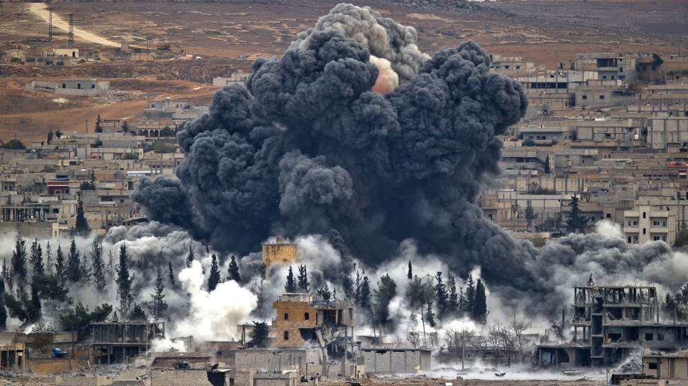 An expansive view of a town experiencing significant devastation with a massive black smoke cloud rising from the ground among residential buildings. The smoke billows aggressively into the sky, obscuring parts of the landscape below, as smaller white plumes of smoke rise around it. The scene is set in a dry, hilly region with sparse vegetation.